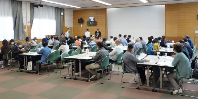 ボランティア連絡会の様子。約60人が、12のグループに分かれてテーブルを向かい合わせにして椅子に座っている。