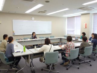 音訳の養成講座の様子の写真。机を円形に並べ、講師が奥に座り、受講者とグループのスタッフ合わせて8名が講師の話を聞いている。