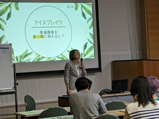 講演会の様子の写真。プロジェクターの前にマイクを持った女性が立っていて、参加者に向けて講義をしている。