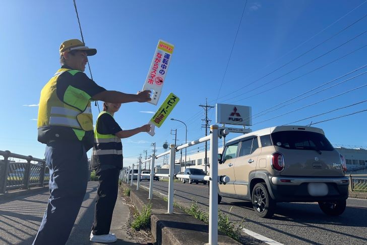 愛知県防犯監視委員会の活動の様子の写真。運転中のドライバーに向けて、歩道から札を振って注意喚起している。札には「スピード注意」や「運転中のながらスマホ禁止」と書かれている。