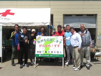 8月活動メンバーの集合写真。
珠洲市災害ボランティアセンターにある「珠洲を応援します　がんばれ！すず　令和7年8月2日(土)」と書かれた看板の前で撮影。男性8人、女性1人。
能登半島の形を倣って右手をグーにして親指を起こして少し曲げた「能登ポーズ」をしている。