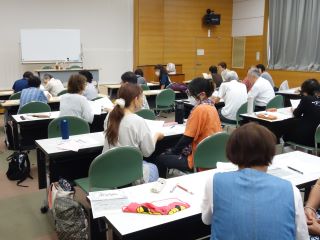 勉強会の様子の写真。参加者が長机1台につき二人掛けで座り、お互い顔を合わせながら話をしている。