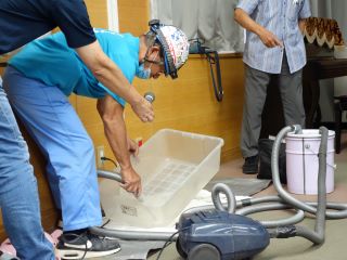 小牧中部地区つどいの写真。チームふじさんの藤野氏が、掃除機の吸い口を持って透明なケースに入れた水を吸い出している。吸い出した水は掃除機からつながったバケツに溜まっている。