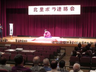 北里地区つどいの写真。ステージ上に岐阜大学の落語研究会の学生芸人のながら家巣春さんが座布団に正座し、扇子を手に落語を披露している。
