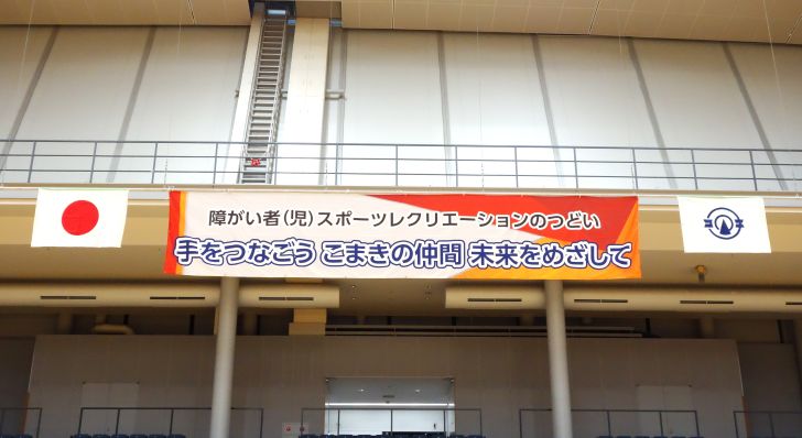 障害者児スポーツレクリエーションのつどいの横断幕が中央にあり、向かって左に日本の国旗、右側に小牧市の市章が掲げられている。