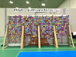 スライム企画の写真。高さ約2m、横約3mのパネルに横板が約8cm間隔で並び、棚板の間に紙コップに入った約500個の色とりどりのスライムが並んでいる。