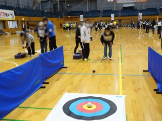 ボッチャ体験の写真。写真手前に得点シートが床に貼られており、3メートルくらい離れて参加者が手のひらより小さいくらいのボールをころがすように投げている。