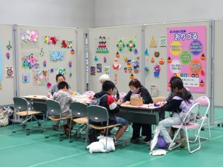 折り紙ボランティア「おりづる」のブースの写真。来場の小学生がボランティアと対面で一緒に折り紙を折っている。