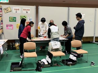 シニア体験グループのブースの写真。高齢者疑似体験中の来場者2名がボランティアから説明を受けている。