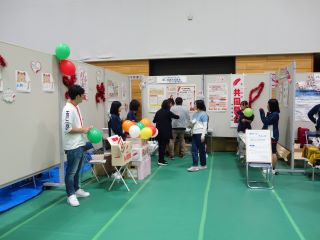 赤い羽根共同募金のブースの写真。パネルに貼られた展示物もあり、来場者は募金をしたり展示物を見たりしている。ジュニア奉仕団の中学生たちもブースを手伝ってくれている。