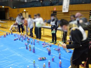 魚つりゲームの様子。ブルーシートが敷かれ、その上には、キラキラひかるモールが中に入った2リットルペットボトルが無造作に置かれている。シートの外から参加者が紐のついた竿についたＳフックでペットボトルの紐にひっかけてつり上げている。