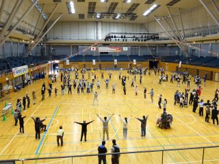 みんなで踊ろうの写真。アリーナの
2階から撮影。30～40人程で輪になって皆さんが両手を上げている。