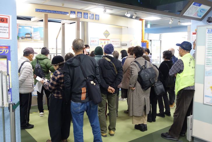 地震体験室の前で集合している写真。20名の参加者が地震体験の前に職員の方の説明を受けている。