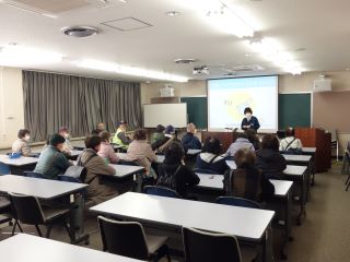 ぼうさい教室の写真。大きい会議室の部屋で、テーブルとイスが並んでいる。机には工作キットのカット済みの牛乳パック、ハサミ、セロハンテープ台が置いてある。