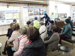 伊勢湾台風の説明を受けている写真。伊勢湾台風の被災した場所の、当時と現在の比較をした写真について、職員の説明を受けている。