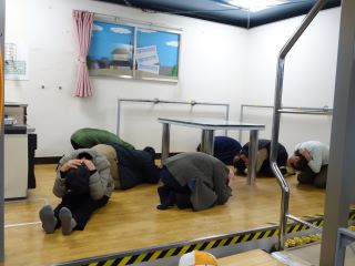 地震体験中の写真。台所を再現した部屋で、部屋の中心にはテーブルが1脚おいてあり、その周りで7名が手で頭や首を押さえて正座になり、うつぶせて揺れに耐えている。