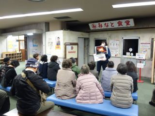 煙避難体験の写真。昭和時代のたたずまいの「みなと食堂」の前で、職員の方から説明を受けている様子。