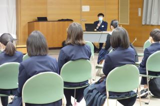 勉強会の写真。手前に椅子に座った中学生の後ろ姿のアップが写っており、その奥に講師の方がパソコンを机に置いて椅子に座って話をしている。