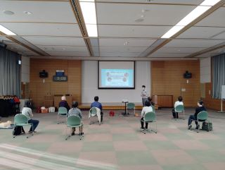 普通救命講習の写真。大会議室に8名の受講生が1～2m間隔で椅子に座り、講師の話を聞いている。