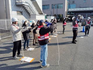 災害ボランティアの実習の写真。十数人が横並びになって、前に立っている講師のボランティアが白いひもを持ち上げて結び方を指導している様子。