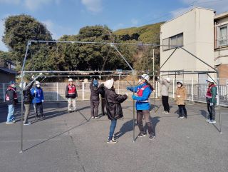 災害ボランティアの実習の写真。ふれあいセンター駐車場で、十数人がテントの骨組みを組み立てている様子。