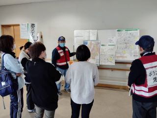 災害ボランティア支援センター演習の写真。事前オリエンテーションの場所で、手前に受講者が並んで、ボランティアの男性がホワイトボードに貼られた資料を見せながら説明をしている様子。