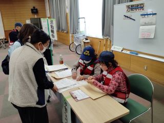 災害ボランティア支援センター演習の写真。ボランティア活動の報告をする場所で、机に並べた資料を座って確認するボランティアの方2名と、その前に立って報告をしている受講者2名の様子。