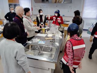 災害ボランティアの実習の写真。料理実習室で講師のボランティアから災害食について説明を受けている様子。