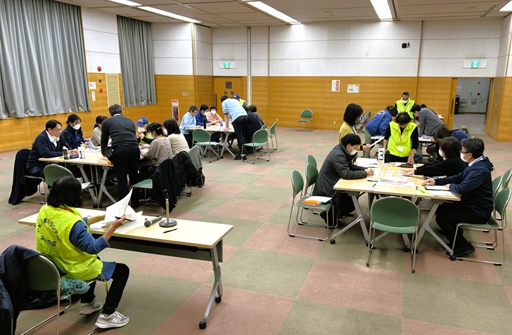 机上訓練1日目の写真。社協職員22名が4班に分かれ、それぞれの班に災害ボランティアネットの会の方が1名ずつ付いてもらっている。長机3つを並べて大きなテーブルにしている。手前には災害ボランティアネットの会の代表が座ってそれぞれの班の様子を見ている。