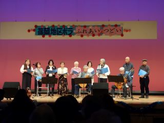つどい舞台発表の写真。2人が椅子に座りギターで伴奏し、9人がその後ろに並んで「いのちの歌」という曲を合唱している。