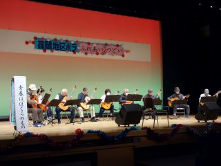 つどい舞台発表の写真。10人弱が椅子に座り、全員がギターを手に「青春はぼくらのもの」という曲を合奏している。