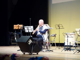 つどい舞台発表の写真。篠岡地区ボランティア連絡会会長の犬飼氏が、ひとり舞台の中心で椅子に座り、尺八を手に演奏している。
