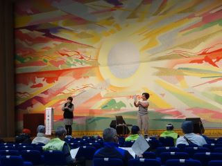 つどい舞台発表の写真。舞台い舞台発表の写真。緞帳の前では、団体の2人が来場者に向かって口腔マッサージの方法などを伝えている。