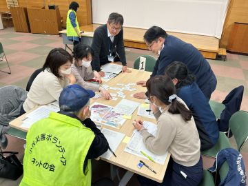 机上訓練1日目の様子の写真。長机3つをつなげ大きなテーブルにし、6名の職員がボランティアカードを並べて話し合っている。手前には災害ボランティアネットの会の方1名がその様子を見守っている。