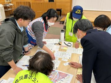 机上訓練2日目の様子の写真。長机3つをつなげ大きなテーブルにし、4名の職員が起立しそれぞれのニーズにどのボランティアを当てるか話し合っている。机の両サイドには災害ボランティアネットの会の方が2名立ち、アドバイスをしている。