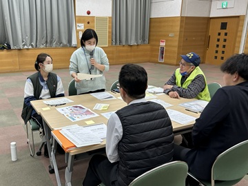 机上訓練2日目の様子の写真。長机3つをつなげ大きなテーブルにし、4名の職員の内1名が起立し話し合いの中で気づいたことなどの発表を行っている。