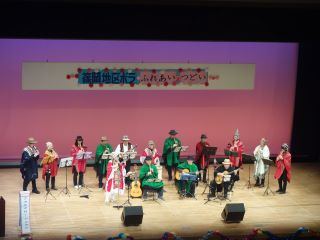 つどい舞台発表の写真。3人が椅子に座り、12人が後ろに並びそれぞれ楽器を持ち、色とりどりのアンデスの衣装に着替えて演奏している。
