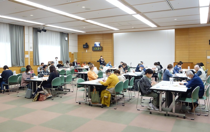 ふれあいセンター大会議室で、約50人が12組に分かれて、ひとテーブル6人掛けで向かい合わせで座っている。