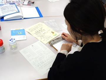 ふれあいセンターボランティアループのテーブルで、参加者の手元を写した写真。点字板と点筆を使い見本を見ながら打っている。