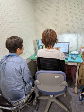 ふれあいセンター録音室に入り録音体験をしている様子。机にパソコンがあり、参加者は椅子に座って録音をし、その様子を「かっこう」の会員の方が見守っている。