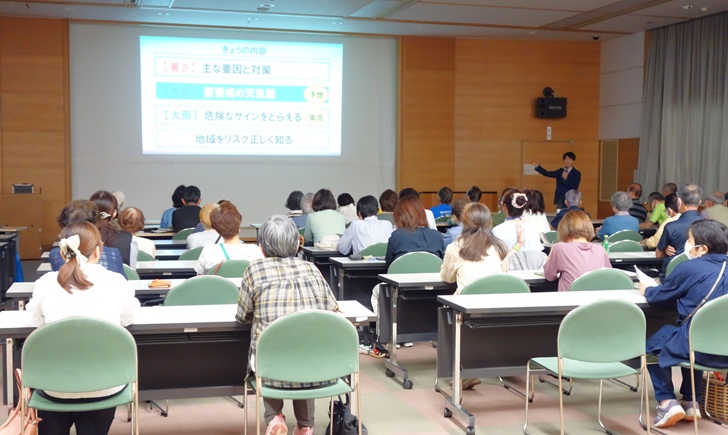 勉強会の写真。参加者の後ろから撮影。画像右奥に講師が立ち、スクリーンに表示されたスライドの説明をしている。「きょうの内容」として、暑さの主な要因と対策や大雨に関することなどの目次が書かれている。