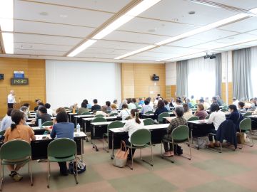 勉強会の写真。ふれあいセンターの3階大会議室で、長机に2人掛けで、横に5つ並び、縦に8つほど並んでいる。74人座れる席に、70名近く座っている様子を後ろから撮影した。