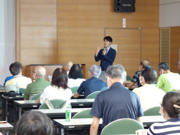 勉強会の写真。手前には参加者の後ろ姿。長机が並び椅子に掛けている。奥には立って話をしている講師。講師はスーツを着ていて、片手でマイクを持ち、参加者の皆さんにむかって笑顔で話をしている。