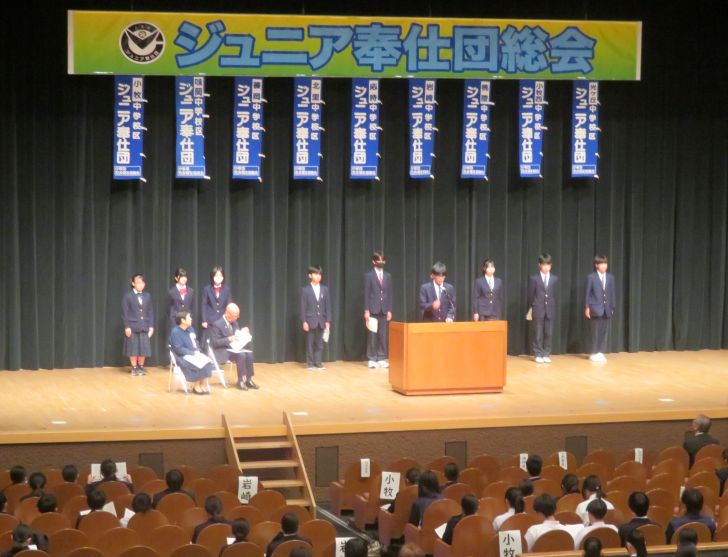ジュニア奉仕団総会の写真。壇上に「ジュニア奉仕団総会」と書かれた横断幕が掲げられている。その下に中学校名が書かれたジュニア奉仕団ののぼり旗が9校分吊り下げられ、それぞれの学校の団員代表はその旗の下に横並びに整列している。向かって左の学校から順番に活動計画発表をしている。社協会長とジュニア奉仕団世話人会副会長が向かって左端にパイプ椅子に座って発表を聴いている。