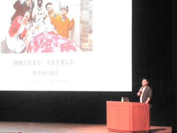 講演の写真。壇上の大きなスクリーンに吉本新喜劇の方々と歌子さん家族が笑顔で写った写真と講演タイトルが映し出されている。右下には講師の関氏。