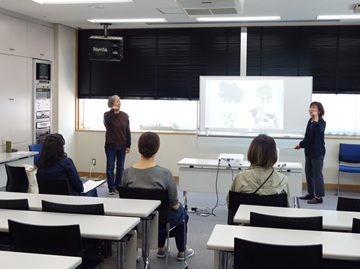手話の養成講座の様子の写真。受講者3名と講師2名が向かい合っている。受講者が椅子に座り横一列に並び、講師はホワイトボードの両脇に立って講義をしている。ホワイトボードにプロジェクターで投影された動物の絵を見ながら、手話での表し方を練習している。