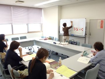 点訳の養成講座の様子の写真。小さめの部屋で、ロの字にテーブルが並んでいる。講師と受講者3名が対面に座り、講師はホワイトボードを使って講義をしている。
