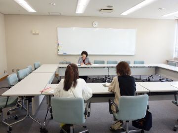 音訳の養成講座の様子の写真。ロの字に並んだテーブルのある部屋で、講師1名と受講者2名が対面に座っている。