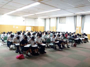 事前説明会の写真。100脚強並んだ椅子に中高生たちが座っている。社協職員から説明を受けながら福祉体験学習用のノートに連絡事項などを書き入れている様子。