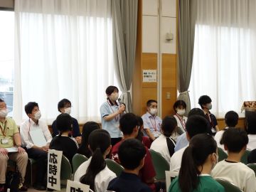 事前説明会の写真。各施設の担当者の紹介。写真奥で窓際に沿って着席している施設担当者が順番に立ち、マイクを手に持って話している。写真手前で座って聞いてる生徒たちに向けて歓迎の言葉をかけている様子。